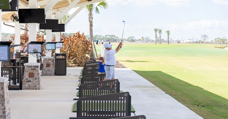 Golfer swinging club at covered driving range with open fairway.