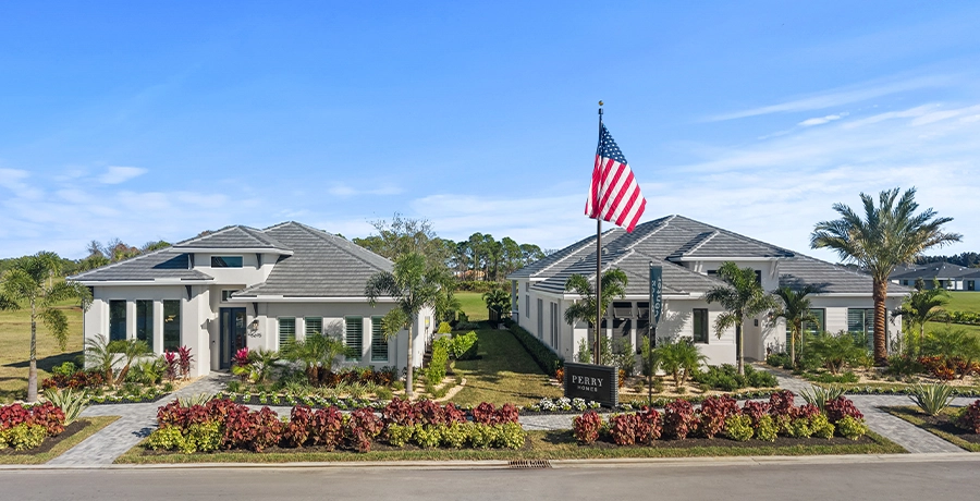 Two modern single-story model homes with landscaped yards, palm trees, and an American flag displayed in front under a blue sky.