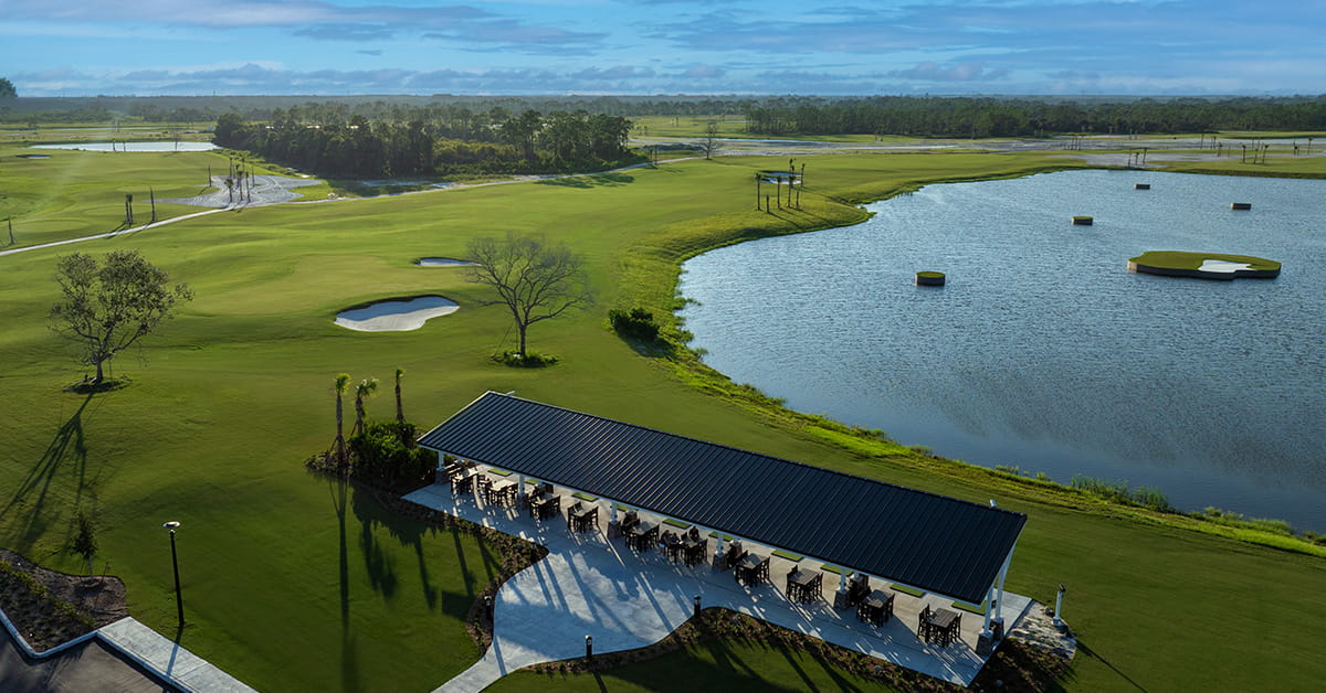 Aerial view of a golf course with a covered pavilion beside a lake, fairways, bunkers, and trees.