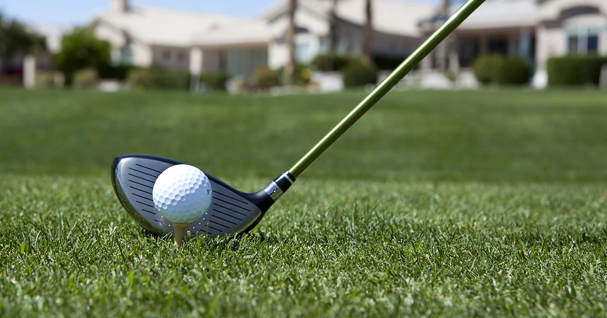 Close-up of a golf ball on a tee against a driver on grass, with a blurred building in the background.