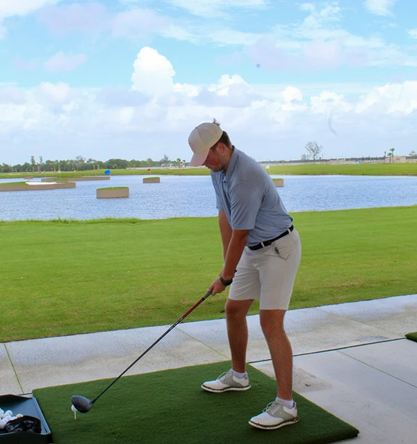 Golfer on a practice mat preparing to hit a teed ball with a driver beside a lake and floating targets.