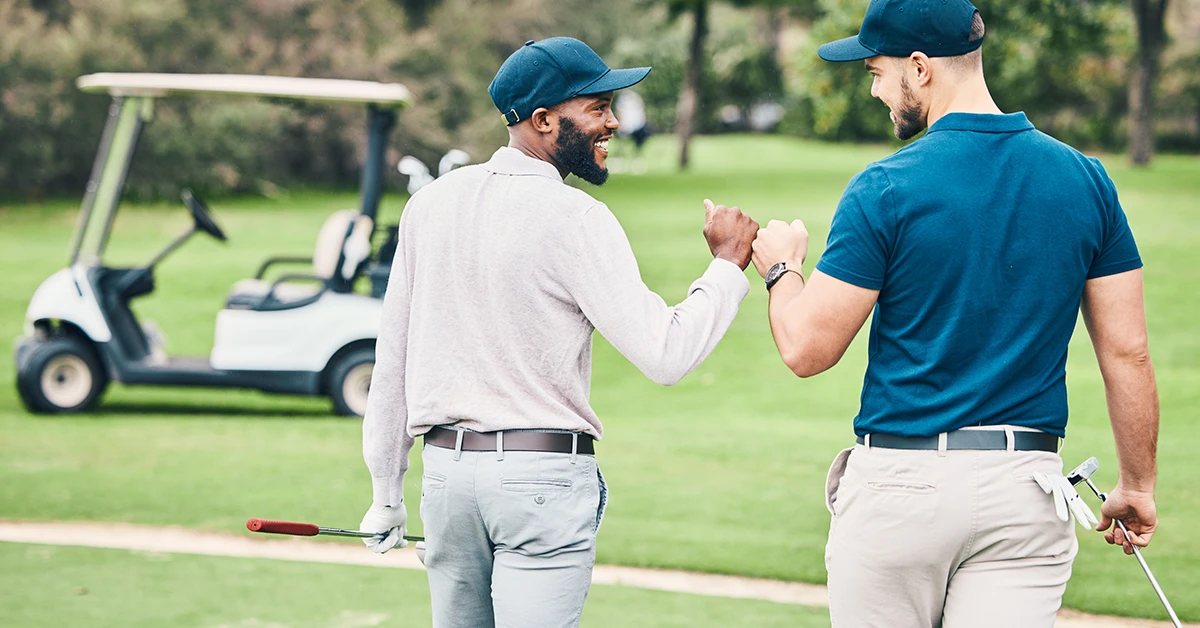 Two golfers bump fists on a golf course while holding clubs, with a golf cart in the background.