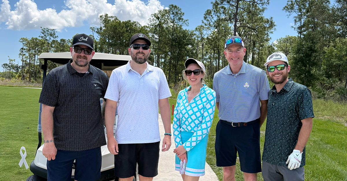 Five golfers pose together beside a golf cart on a golf course.