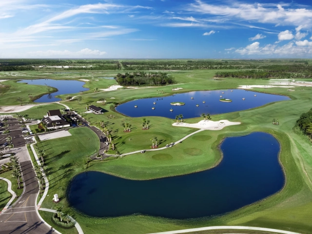 Aerial view of a clubhouse building next to a pond with floating golf driving range targets, palm trees, and a parking lot.
