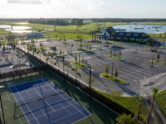 Aerial view at golden hour of a clubhouse, sport courts, parking lot, and a pond with floating golf targets, surrounded by homes.