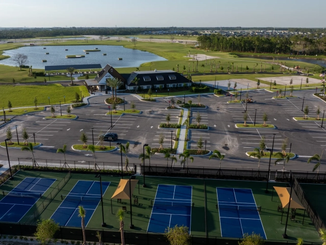 Aerial view at golden hour of pickleball courts, a clubhouse, parking lot, and a pond with floating golf targets, with homes in the distance.