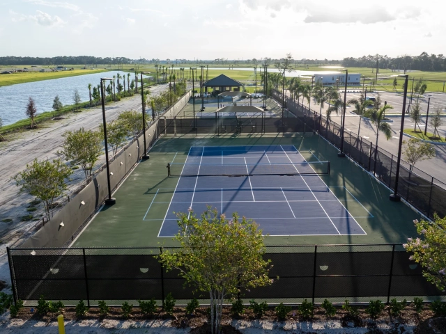 Aerial close-up of a fenced tennis or pickleball court next to a lake, with a parking lot and palm trees nearby.