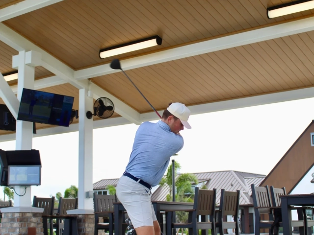 A man mid-golf-swing inside a covered driving range bay with wood ceilings and wall-mounted TVs showing golf content.