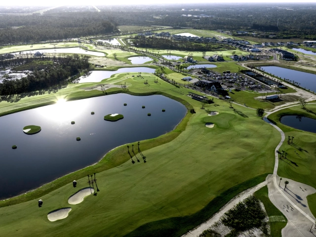 Wide aerial view of a golf course community with a clubhouse, driving range pond, multiple ponds, and homes.