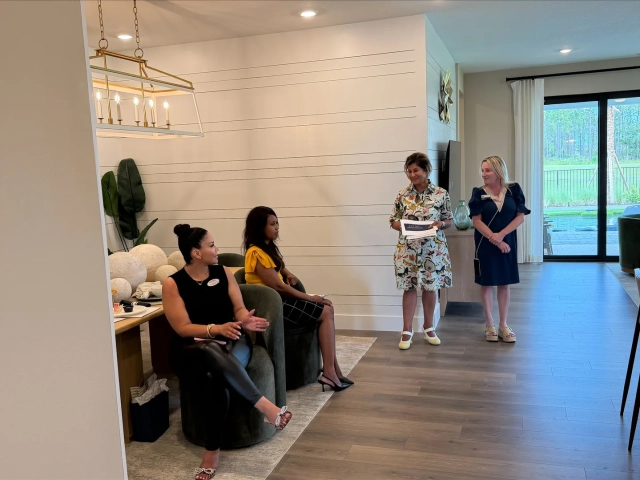 Two women standing and presenting a prize card to two seated women in a model home living room.