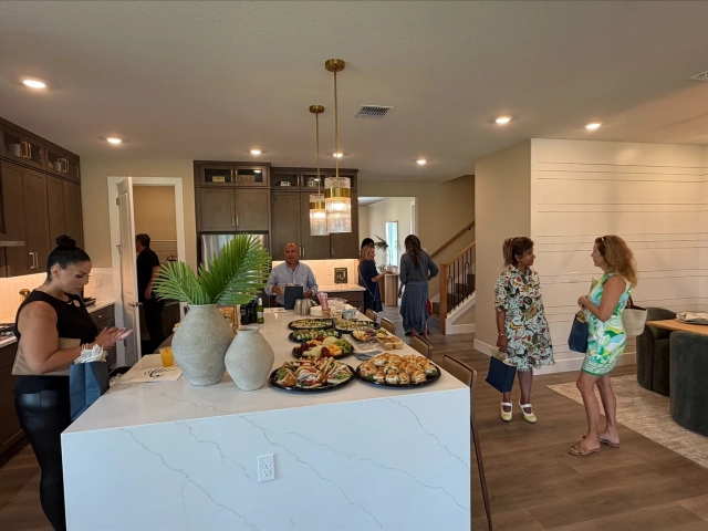Guests standing and talking near a kitchen island topped with trays of sandwiches, fruit, and pastries.