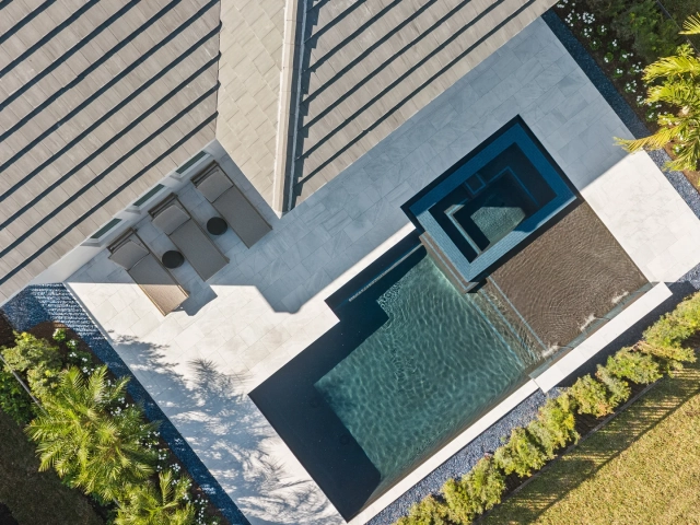 Aerial view of a geometric backyard pool with a raised spa, lounge chairs, and paver patio surrounded by palm trees.