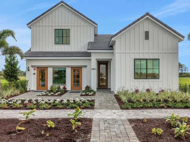 Two-story white farmhouse-style model home with dark roof, wood-paneled entry doors, and landscaped front walkway, address 6671.