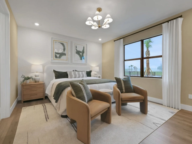 Primary bedroom with a white upholstered bed, two tan leather chairs, and a large window overlooking a golf course.